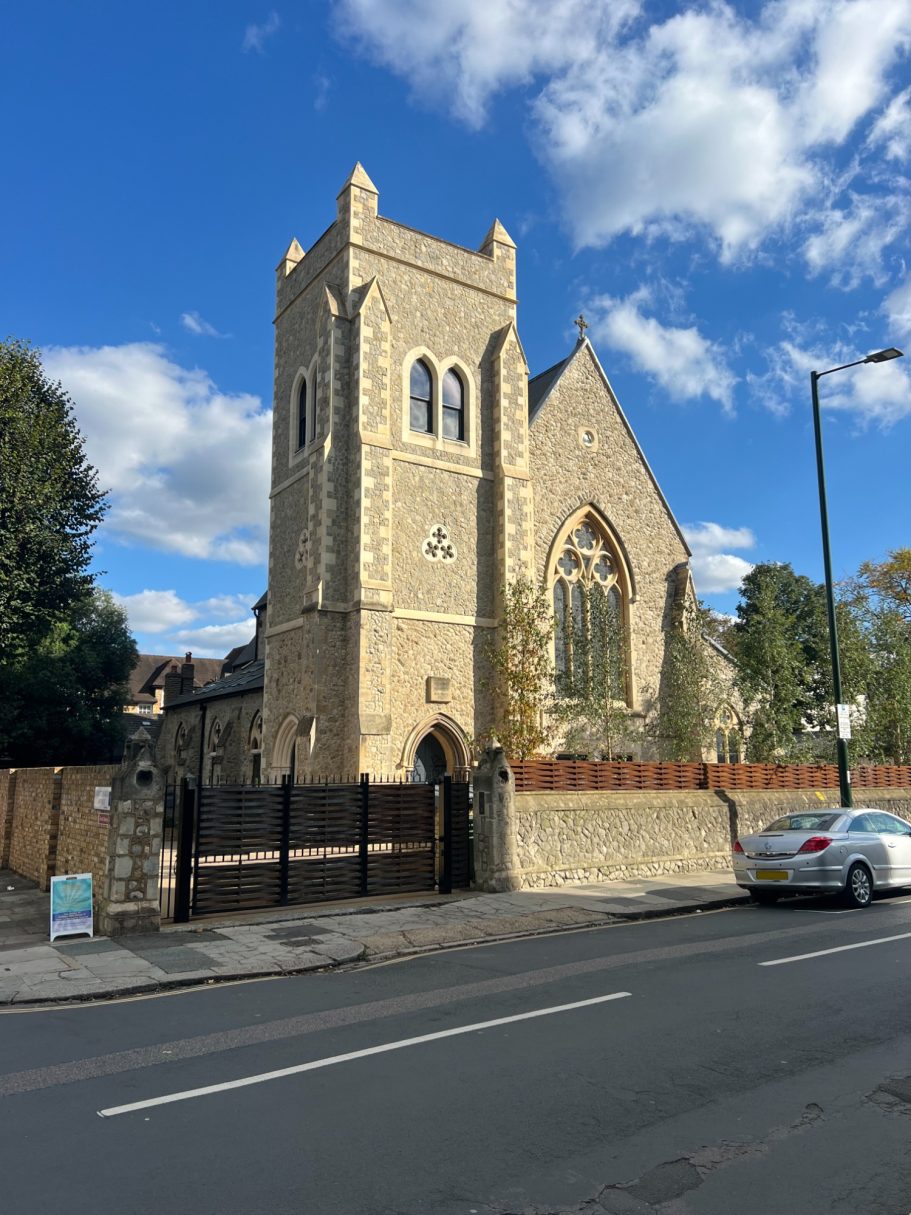 Victorian-style stone church with a tall tower, set against a blue sky and trees.