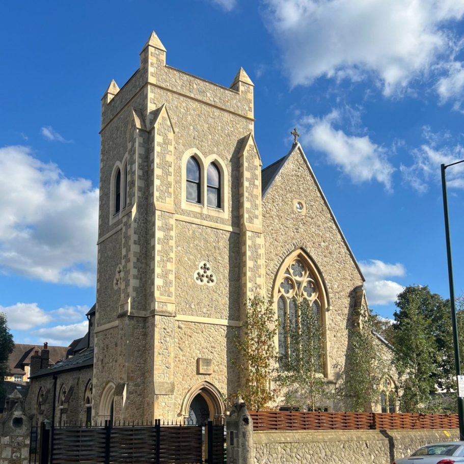 Stone church with a tall tower, surrounded by trees under a blue sky with clouds.